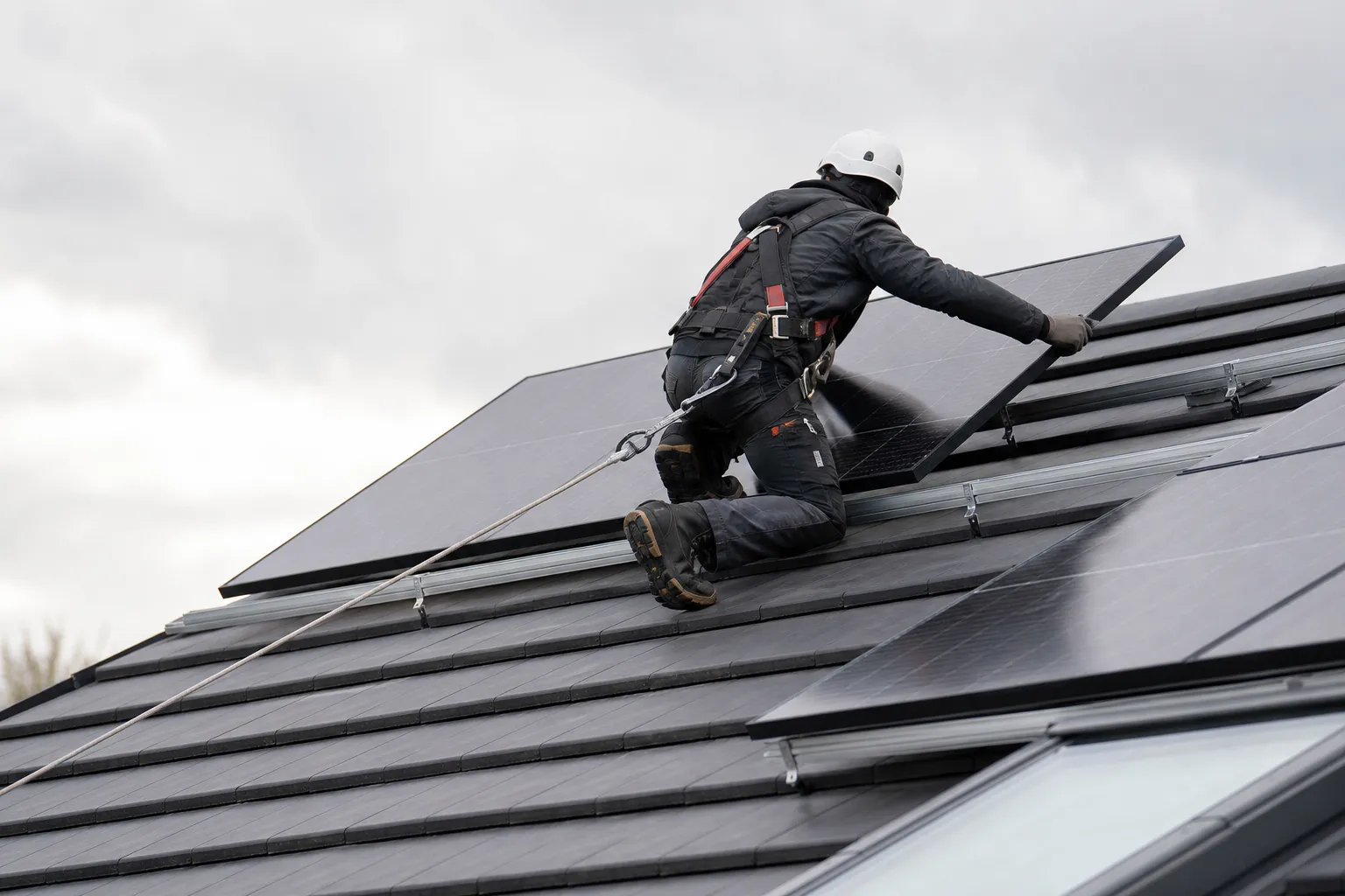 Technicien harnaché posant un panneau solaire sur une toiture en pente, vue de dos